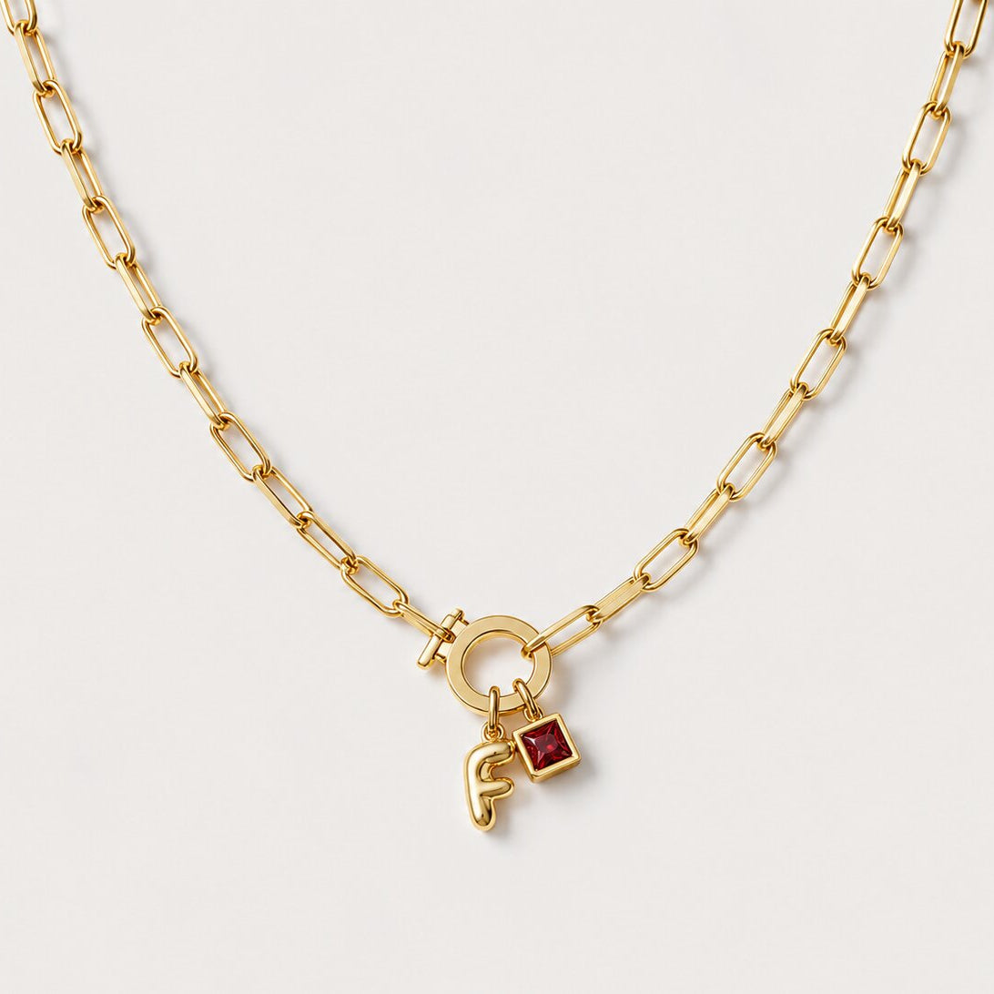 A gold chain necklace featuring a small padlock charm with a red birthstone gem and a gold monogram pendant, displayed on a clean off-white background.