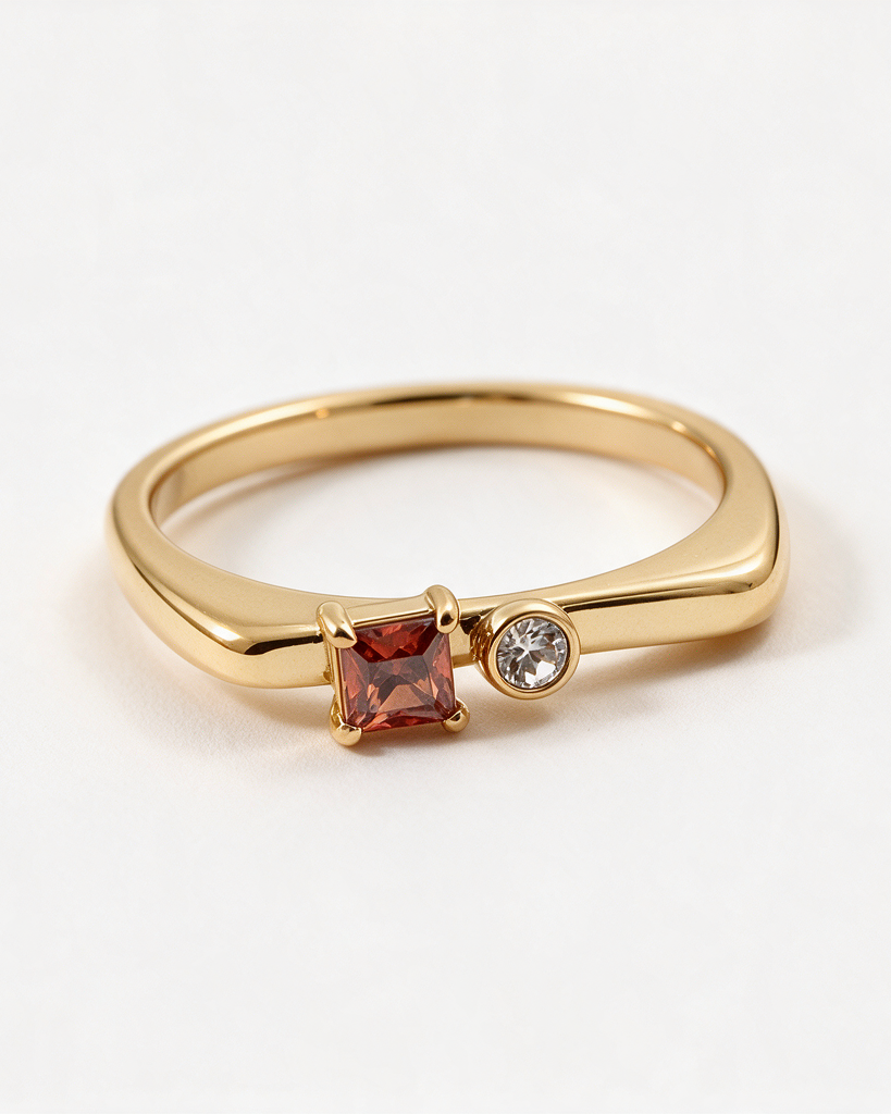 A gold band ring featuring a princess-cut red gemstone and a small round clear stone, photographed on a clean white background.