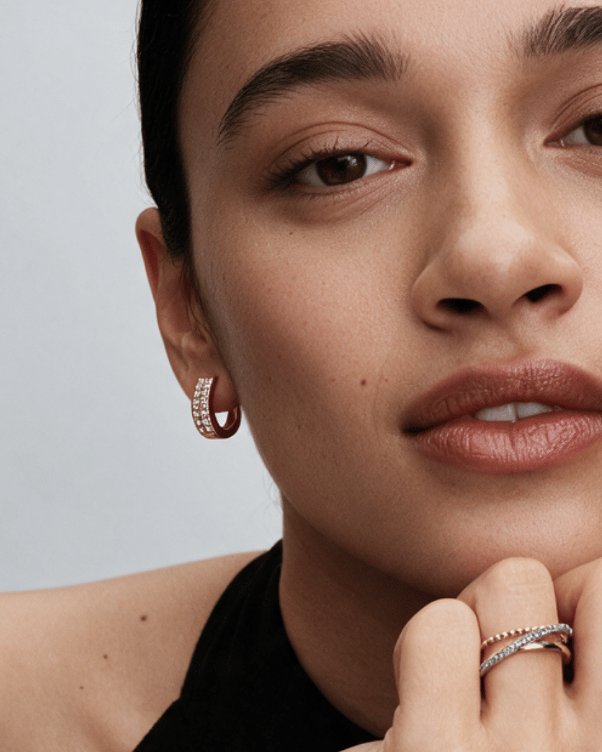 Close-up of a model wearing a small hoop earring with pavé stones and stacked rings on her fingers, set against a clean light gray background.