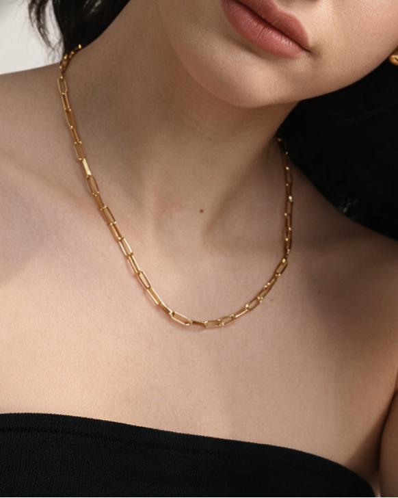 A close-up lifestyle shot of a woman wearing a gold chain necklace, matching gold stud earrings, and a gold link bracelet, paired with a black strapless top.