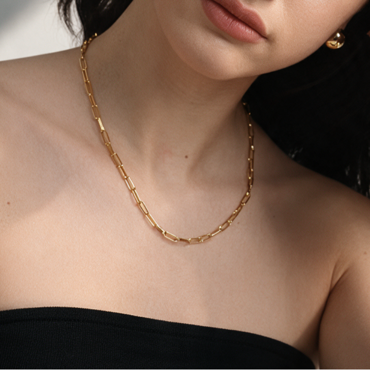 A close-up lifestyle shot of a woman wearing a gold chain necklace, matching gold stud earrings, and a gold link bracelet, paired with a black strapless top.