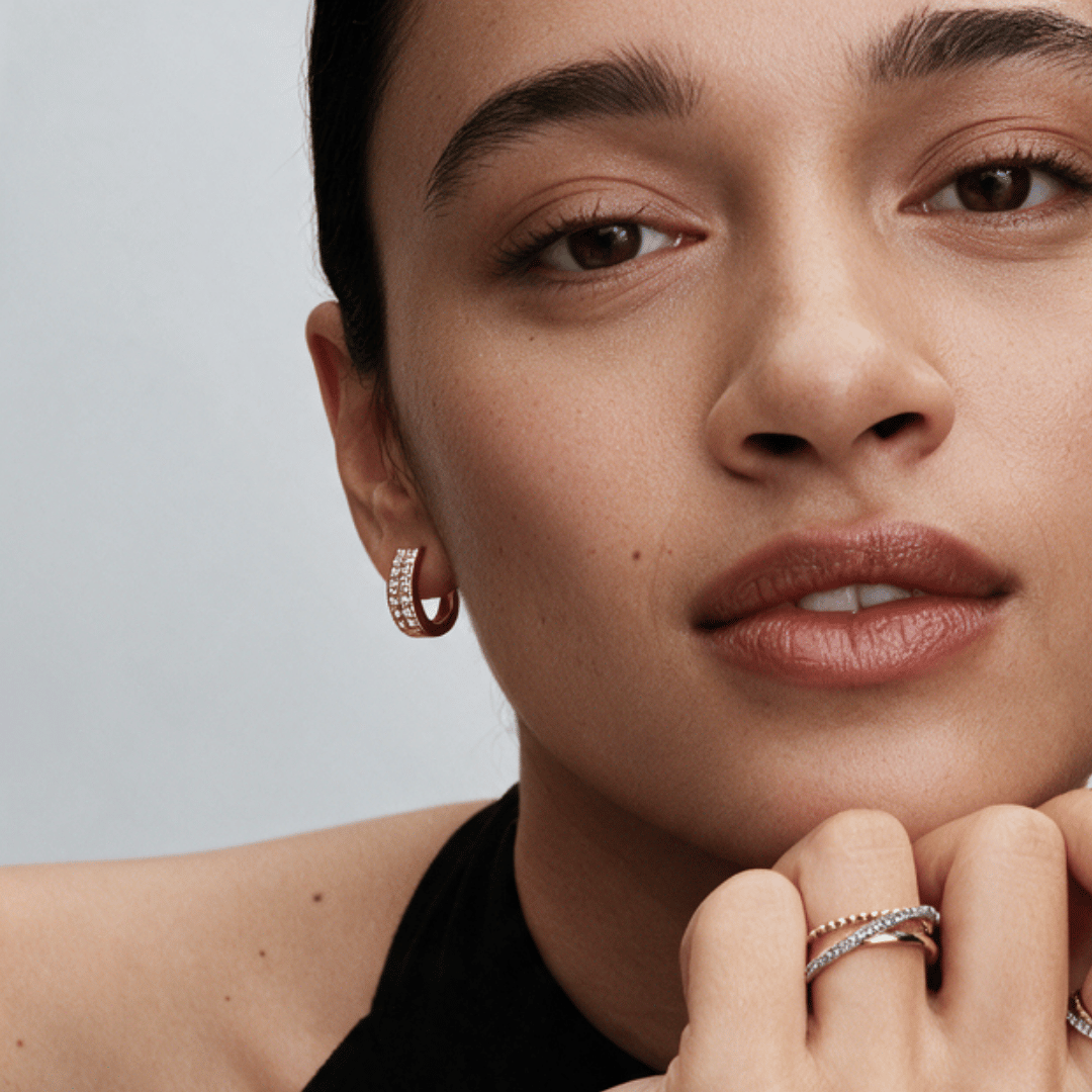Close-up of a model wearing a small hoop earring with pavé stones and stacked rings on her fingers, set against a clean light gray background.