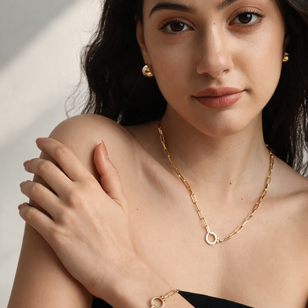 A close-up lifestyle shot of a woman wearing a gold chain necklace with a circular ring pendant, matching gold stud earrings, and a gold bracelet.